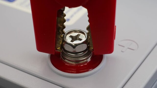 Red alligator clip grips positive battery terminal with metal teeth. Close-up shows secure electrical connection for battery charging or jump-starting process