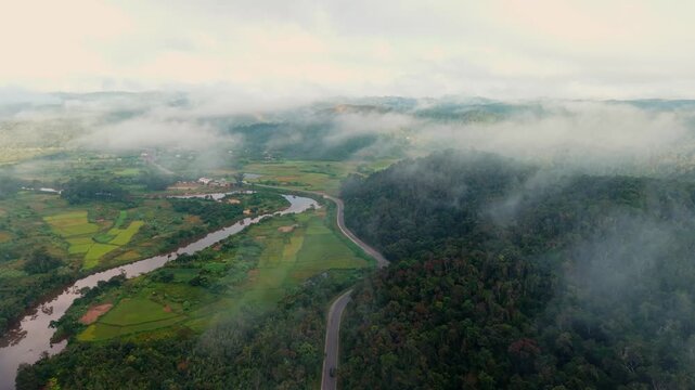 This stunning aerial drone shot follows the brown river as it winds past a small jungle village and green rice fields tucked into the misty mountains of rural Madagascar