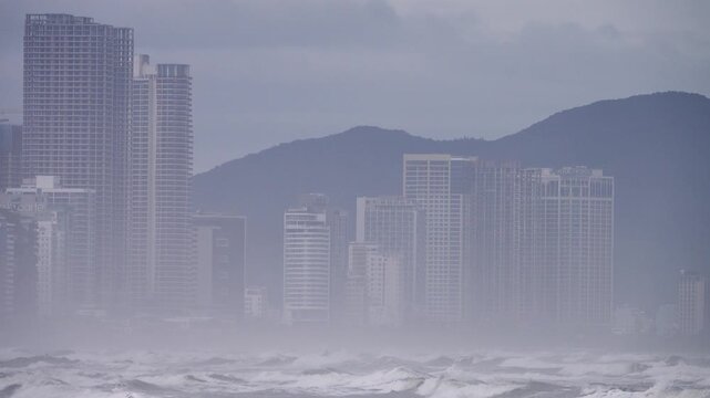 The South China Sea during the calendar winter and seasonal transition, rainy season, northeast winter monsoon, blowing spray. Bay is covered with foam waves and hotel buildings on shore. Vietnam