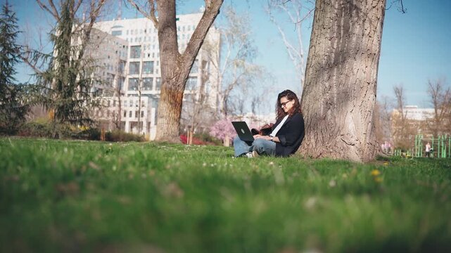 Young student woman in glasses working on a laptop under a tree in a blooming spring park. Outdoor studying, education, focus, and modern student lifestyle concept.