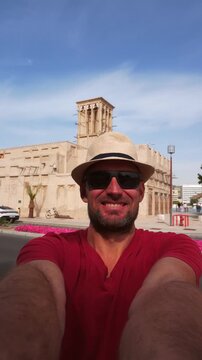 A happy tourist wear red shirt and straw hat takes selfie at the Al Fahidi Historical Neighborhood in Old Dubai. Features a traditional wind tower backdrop cityscape view
