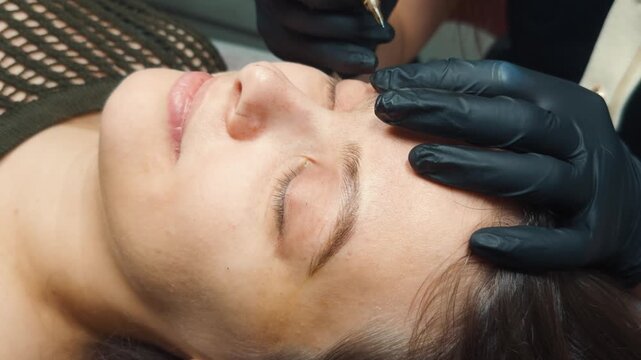 Woman receiving microblading permanent makeup treatment, beauty professional applying cosmetic pigment creating natural hair strokes on eyebrows with specialized tool
