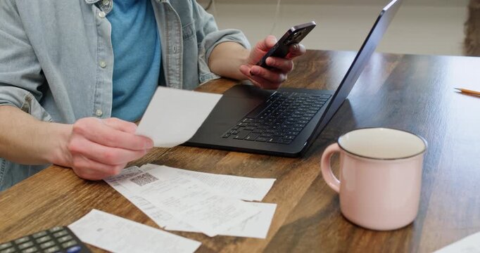 Man checks paper receipt while using smartphone near laptop and tea mug. Person compares payment details on documents managing household expenses at table slow motion