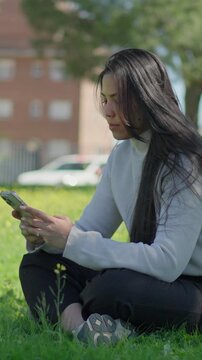 Young woman texting on smartphone while sitting on green grass in city park