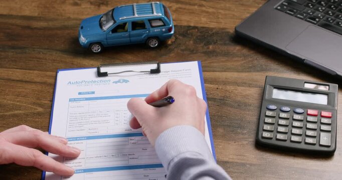 Driver writes on vehicle report form near toy car and calculator on desk. Man reviews insurance details filling policy fields with hand during claim process slow motion