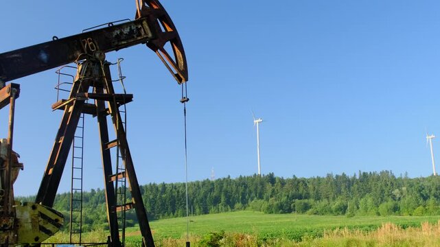 Old oil well with rusty pumpjack stand against wind turbines on hill slow motion. Transition to clean renewable power sources at industrial farm
