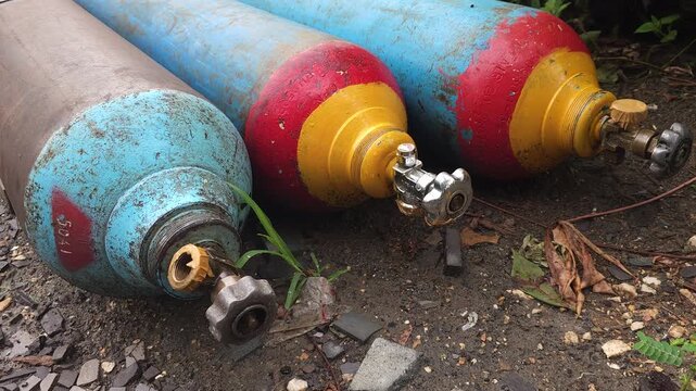Weathered and rusted compressed gas cylinders are left abandoned on rough outdoor ground surrounded by gravel and weeds. The industrial metal tanks, equipped with heavy-duty valves