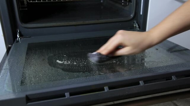 Person cleaning oven glass door with damp sponge, applying cleaning solution to surface, diligent effort for spotless kitchen appliance and improved home hygiene