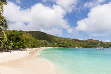 Obraz premium A tranquil tropical beach with powder-white sand, clear turquoise water, swaying palm trees and an anchored yacht framed by green hills and a bright blue sky with soft white clouds. Seychelles