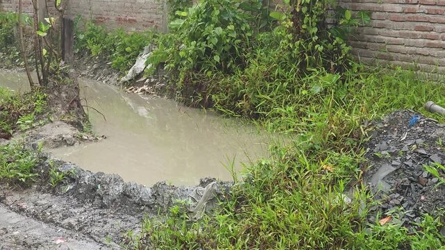 Murky, brown water flows slowly through a muddy drainage ditch surrounded by overgrown weeds and tall grass. A pile of rubble and a plastic pipe sit on the muddy bank