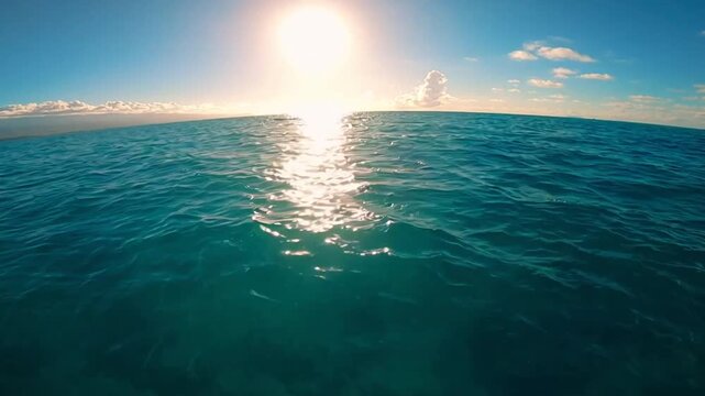 Bright sun shining over a deep blue ocean with sparkling water reflections and small ripples under a clear sky with scattered white clouds on the horizon at sea level.