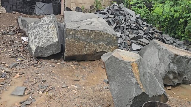 Large, unpolished stone boulders and piles of broken rock fragments sit outside a small-scale, informal stone processing workshop. The rough exterior of the building houses basic machinery