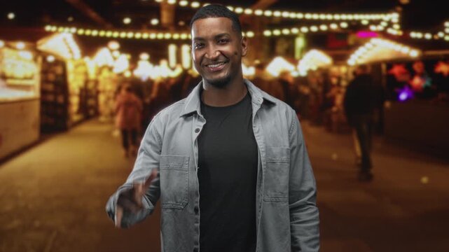 Young black man smiling and making i love you hand sign on a street lined with market stalls and string lights; joyful connection.