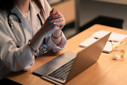 A medical professional in a white coat with a stethoscope sits at a desk with a laptop, notebook, and glass of water, working late into the night.