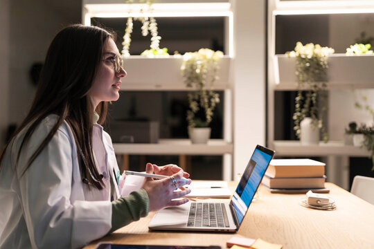 A medical professional in a lab coat works late at a desk with a laptop and tablet, reviewing patient information.