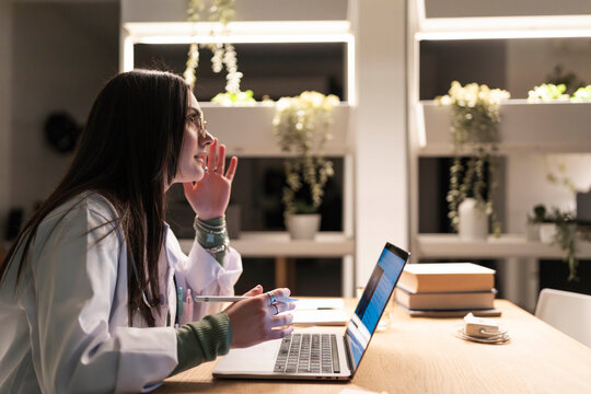 A medical professional in a lab coat works late at a laptop, holding a stylus and looking thoughtfully at the screen.
