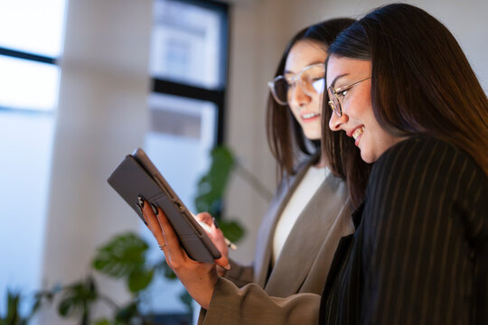Two women in glasses collaborate on a digital tablet, discussing ideas and working together in a modern office setting.