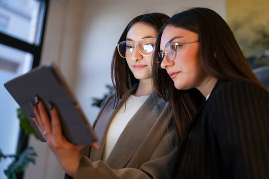 Two young women wearing glasses collaborate on a digital tablet, sharing ideas and information in a modern office setting.