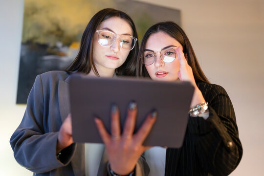 Two young women wearing glasses collaborate on a digital tablet, their faces illuminated by the screen's glow as they focus on the content.