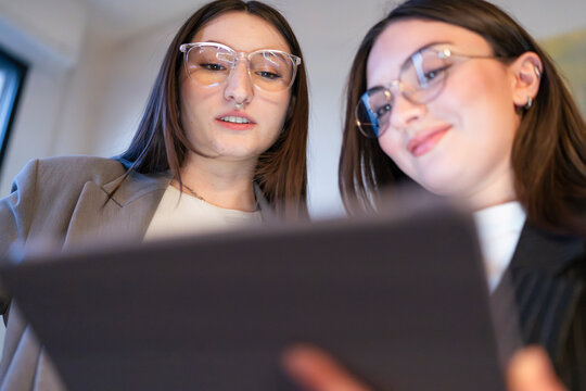 Two young women wearing glasses collaborate over a digital tablet, discussing a project in a modern office setting.