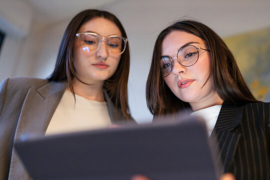 Two young women in business attire collaborate over a digital tablet, focusing intently on the screen.
