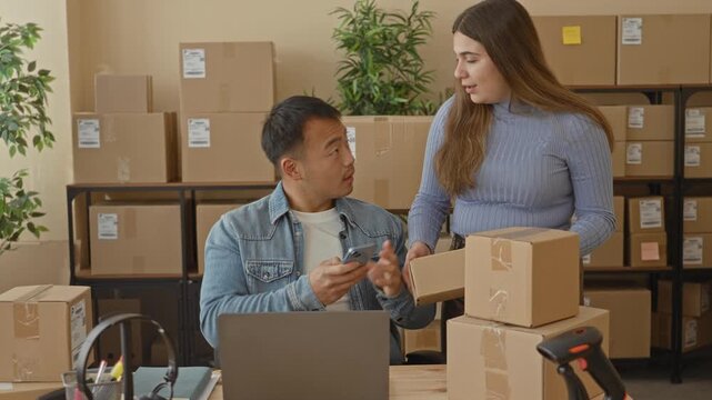 Man points smartphone at cardboard box while woman hands over a parcel and gestures toward stacked boxes in building; teamwork focused.