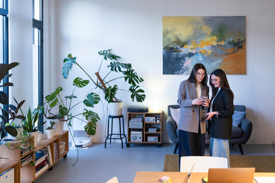 Two women in business attire stand together, looking at a smartphone. They are in a modern office space with plants and artwork.