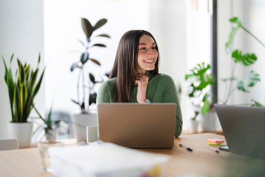 A young woman smiles while working on her laptop in a bright, modern office space with plants.