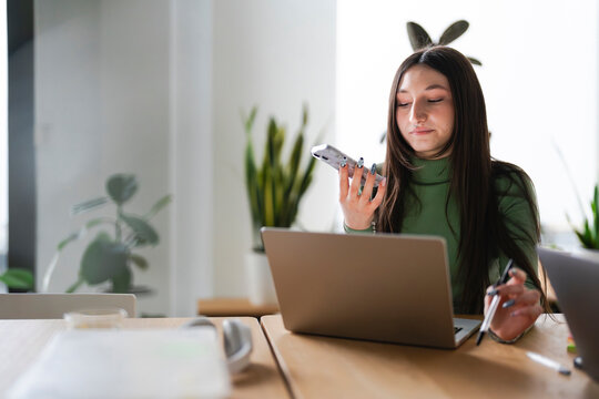 A young woman works on her laptop while holding a phone and a pen, focused on her tasks in a bright, modern office setting.