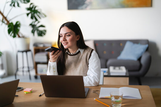 A young woman is working from home, using her phone to record a voice message while sitting at her desk with a laptop and notebook.