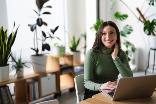 A young woman smiles while working on her laptop in a bright, plant-filled room. She is wearing a green turtleneck and has her hand on her chin.