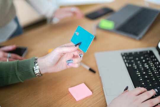 A person holds a debit card while typing on a laptop, surrounded by office supplies, suggesting online shopping or financial transactions.