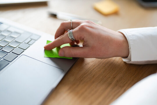 A person's hand with rings touches a green sticky note with writing on it, placed on a laptop keyboard.