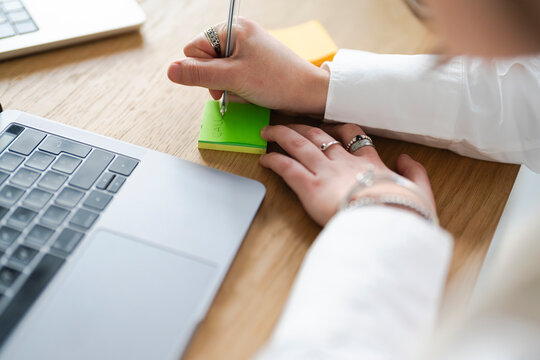 A person's hands are writing on a sticky note next to a laptop, suggesting a moment of planning or task management.