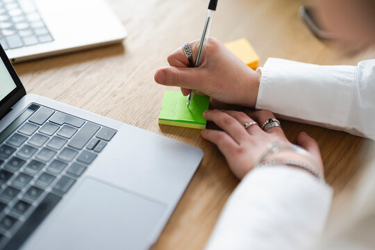 A person writes a note on a sticky pad next to a laptop, indicating a task to be done.
