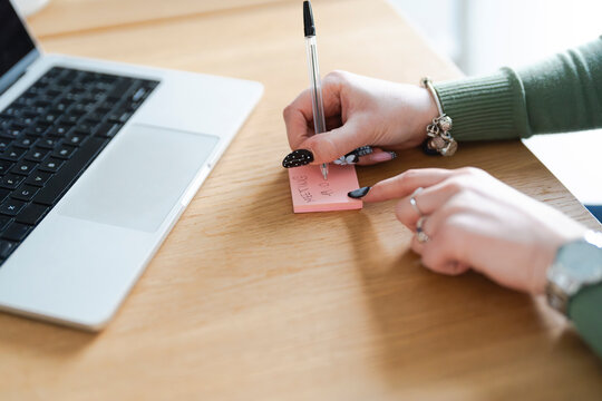 A person's hands write a note on a pink sticky pad next to a laptop, indicating a meeting time.