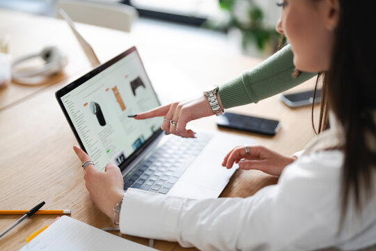 Two women collaborate on a laptop, pointing at the screen displaying an online shopping interface, suggesting teamwork and digital commerce.