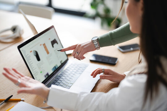 Two women collaborate on a laptop, one pointing at the screen displaying an online shopping page, suggesting teamwork and digital commerce.