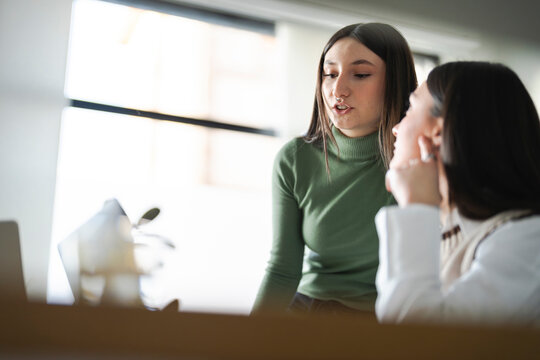 Two young women are engaged in a conversation, possibly discussing a project or idea, in a bright, modern office setting.
