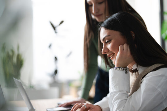 Two young women collaborate on a project, one looking at a laptop screen while the other leans in to observe.