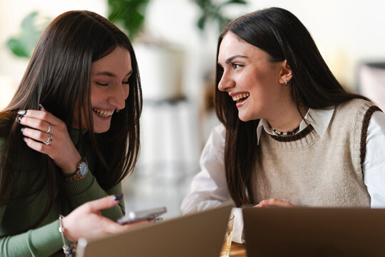 Two young women laugh while looking at a mobile phone, sharing a moment of joy and connection over a table with laptops.