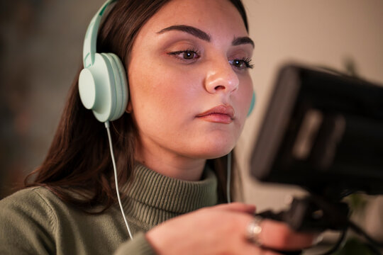 A young woman wearing headphones and a green turtleneck is focused on a screen, holding a device in her hand.