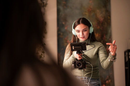 A young woman wearing headphones operates a video camera with a monitor attached, gesturing with her hand as she records.