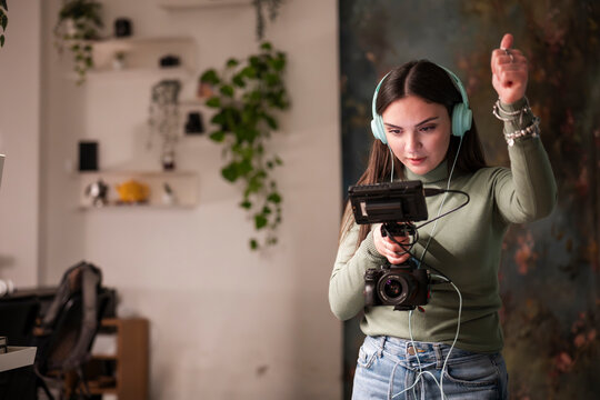 A young woman wearing headphones operates a camera with a monitor, her hand raised in a gesture of direction or approval.