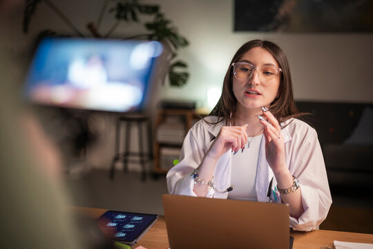 A young woman in a lab coat and glasses is speaking while looking at a laptop and tablet displaying medical images.