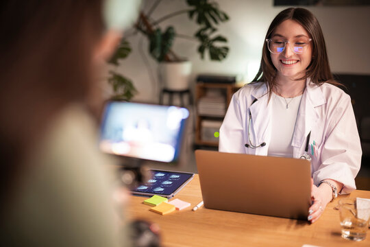 A smiling doctor in a white coat consults with a patient, reviewing medical images on a tablet and laptop during a telehealth appointment.