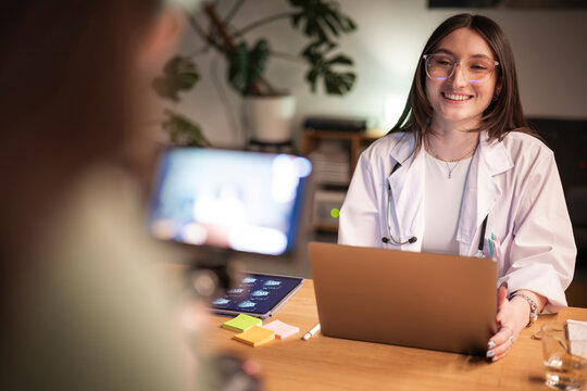 A smiling doctor in a white coat with a stethoscope consults with a patient via video call on a laptop, reviewing medical images on a tablet.