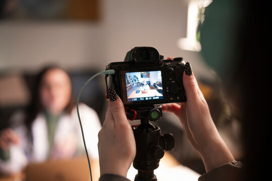 A person films another person in a room using a digital camera on a tripod. The camera screen shows the subject being filmed.