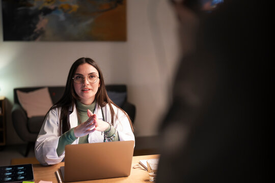A female doctor in a white coat and glasses consults with a patient, using a laptop and tablet in a modern office setting.