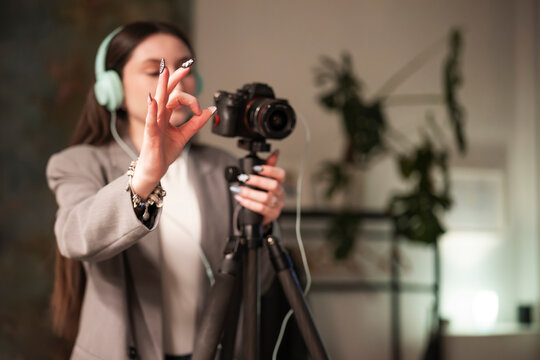 A woman wearing headphones and a blazer is filming with a camera on a tripod, making an okay gesture with her hand.